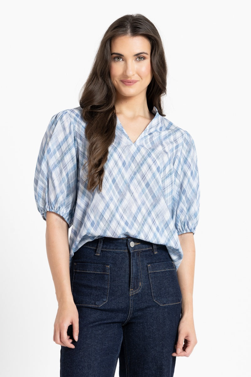 A woman modeling a light blue and white plaid short-sleeve blouse with puff sleeves and a V-neckline, paired with dark blue jeans, against a plain white studio background.