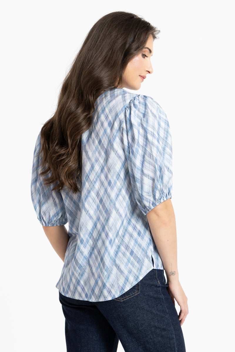 A woman shown from the back wearing a light blue and white plaid puff-sleeve blouse with dark blue jeans, photographed against a plain white studio background.
