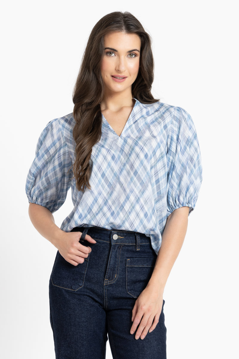 A woman modeling a light blue and white checkered puff-sleeve blouse with a V-neckline, paired with dark blue jeans, against a plain white studio background.