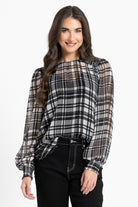 A woman modeling a black, white, and gray plaid blouse with sheer long sleeves and a gathered neckline, paired with black pants, against a plain white background.