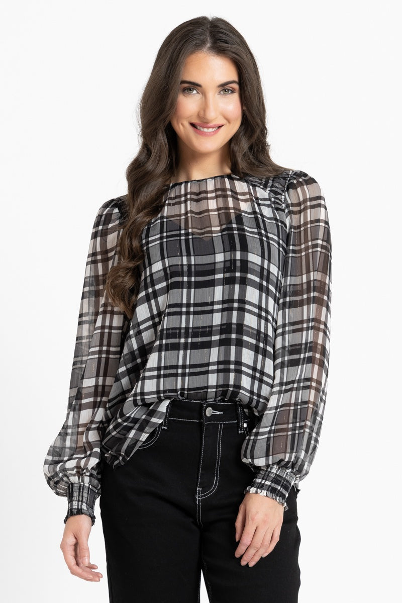 A woman modeling a black, white, and gray plaid blouse with sheer long sleeves and a gathered neckline, paired with black pants, against a plain white background.