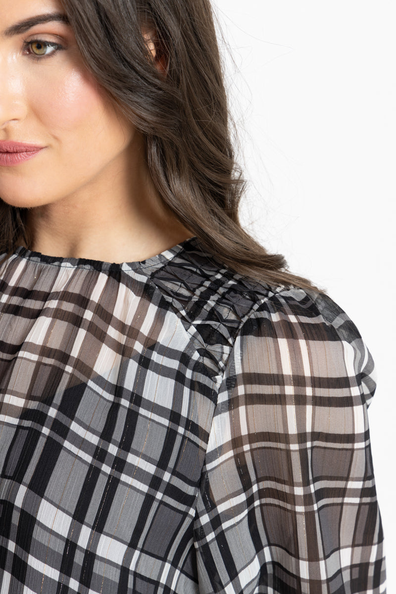 Close-up of a woman wearing a sheer black, white, and gray plaid top with textured shoulder detailing, shown against a plain white background.