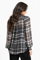 A woman shown from the back wearing a sheer long-sleeve plaid blouse in black, white, and brown tones, paired with black pants, against a plain white studio background.