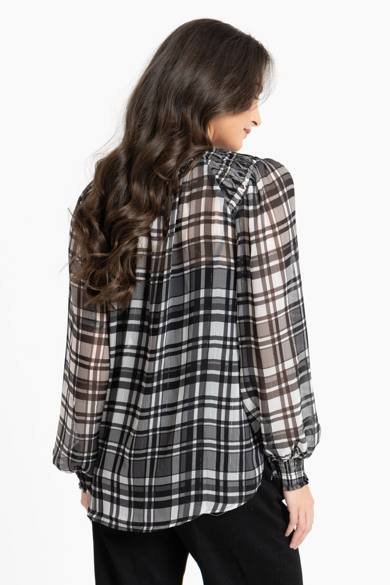 A woman shown from the back wearing a sheer long-sleeve plaid blouse in black, white, and brown tones, paired with black pants, against a plain white studio background.