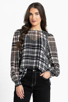A woman wearing a black, white, and gray plaid blouse with sheer long sleeves and puff sleeves, paired with black pants, against a plain white studio background.