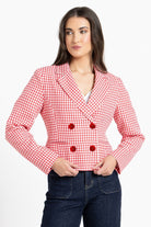 A woman modeling a red and white gingham double-breasted blazer with red buttons, paired with dark blue jeans, against a plain white background.