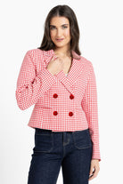 A woman modeling a red and white gingham double-breasted blazer with red buttons, paired with dark blue jeans, against a plain white background.