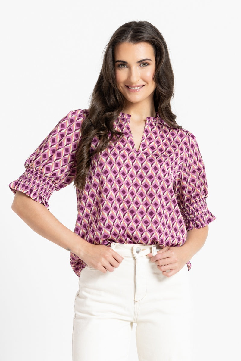 A woman modeling a short-sleeve blouse with a purple and pink geometric diamond pattern, paired with white pants, against a plain white background.