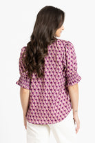 A woman viewed from the back wearing a pink and white patterned blouse with puff sleeves and white pants, set against a plain white background.