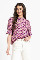 A woman modeling a short-sleeve blouse with a purple, pink, and white geometric pattern, paired with white pants, against a plain white background.