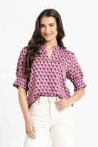 A woman modeling a short-sleeve blouse with a purple and white geometric pattern, paired with white pants, against a plain white background.