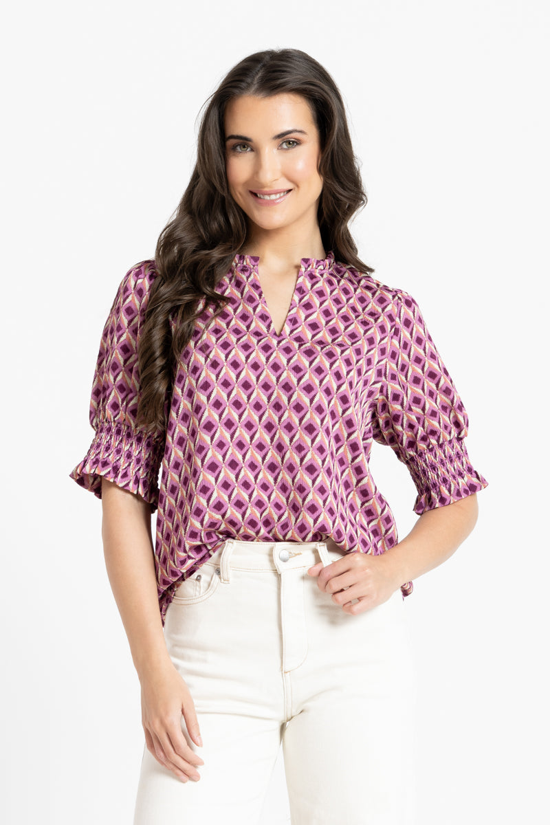 A woman modeling a short-sleeve blouse with a purple and white geometric pattern, paired with white pants, against a plain white background.