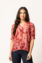 A woman modeling a short-sleeve button-up blouse with a dusty rose and coral floral print, paired with dark blue jeans, against a plain white studio background.