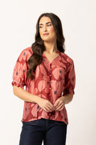 A woman modeling a short-sleeve button-up blouse with a dusty rose and red floral pattern, paired with dark blue jeans, against a plain off-white background.