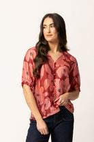 A woman modeling a dusty rose pink short-sleeve button-up blouse with a dark floral pattern, paired with dark blue jeans, against a plain white studio background.