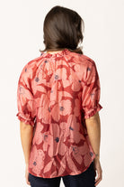 A woman viewed from the back wearing a coral pink short-sleeve blouse with a dark floral print and ruffled cuffs, paired with dark jeans against a plain white background.