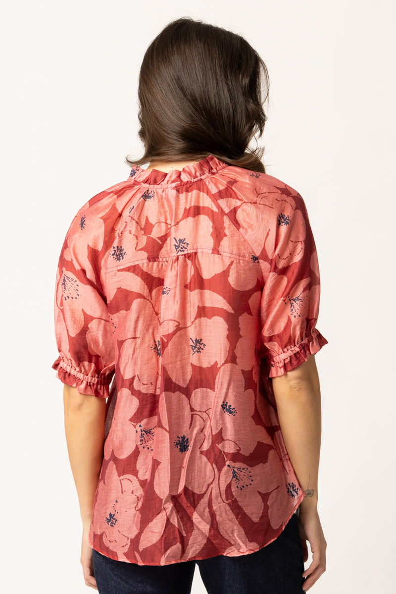 A woman viewed from the back wearing a coral pink short-sleeve blouse with a dark floral print and ruffled cuffs, paired with dark jeans against a plain white background.