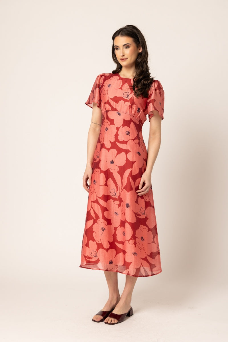 A woman modeling a red midi dress with a large pink floral pattern, short flutter sleeves, and a round neckline, standing against a plain beige studio background.