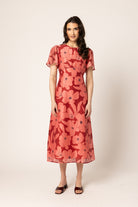 A woman modeling a short-sleeved midi dress with a red and coral floral pattern, standing against a plain beige studio background.