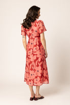 A woman is shown from the back wearing a coral red midi dress with a large floral print in pink and orange, paired with dark brown flat shoes, against a plain cream background.