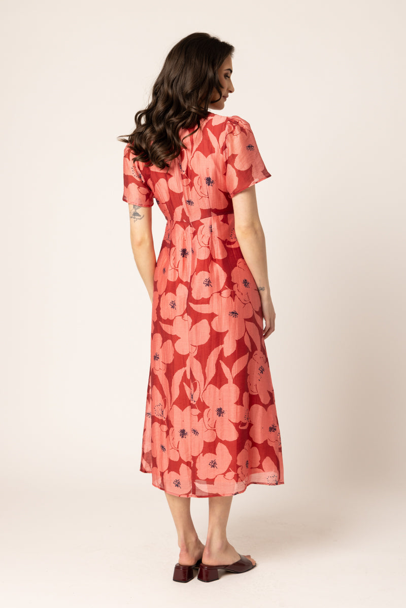 A woman is shown from the back wearing a coral red midi dress with a large floral print in pink and orange, paired with dark brown flat shoes, against a plain cream background.