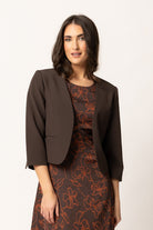 A woman modeling a dark brown cropped blazer layered over a brown dress with an orange floral pattern, set against a plain off-white studio background.
