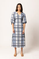A woman modeling a blue and white plaid midi dress with puff sleeves and a V-neckline, standing against a plain light beige studio background.