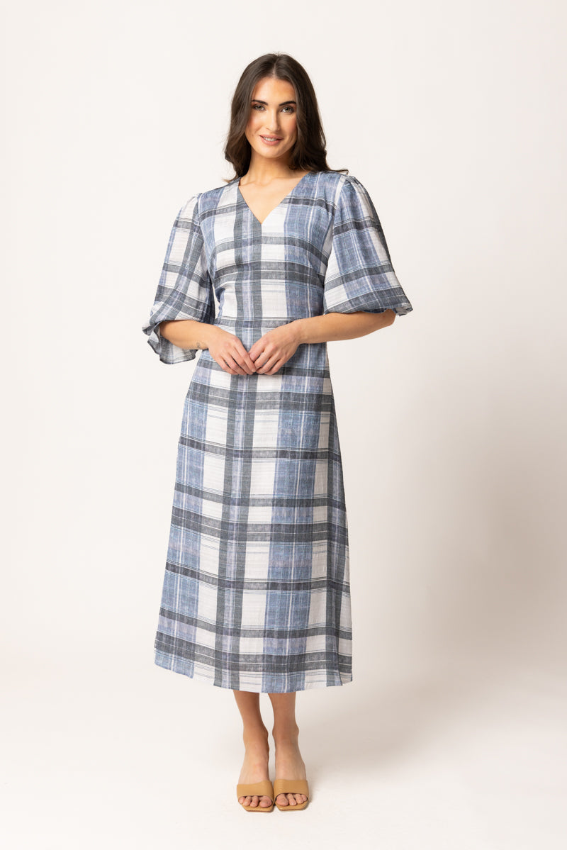 A woman modeling a blue and white plaid midi dress with elbow-length flutter sleeves and a V-neckline, standing against a plain light beige studio background.