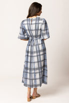 A woman viewed from the back wearing a midi dress with a blue, gray, and white plaid pattern, puff sleeves, and a tie waist, paired with tan flat sandals against a light beige studio background.