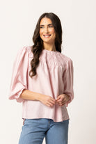 A woman with long dark hair wearing a light pink smocked blouse with puff sleeves and blue jeans, standing against a plain off-white studio background.