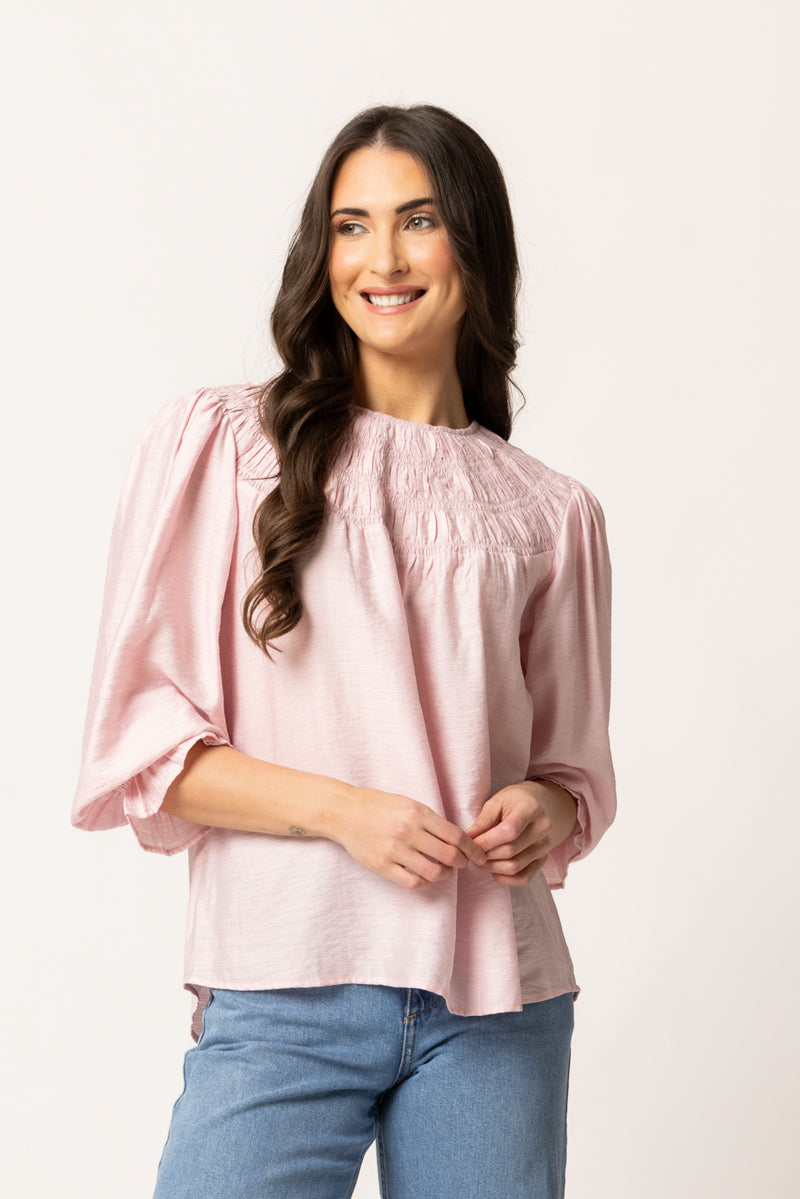 A woman with long dark hair wearing a light pink smocked blouse with puff sleeves and blue jeans, standing against a plain off-white studio background.