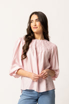 A woman with long dark wavy hair wearing a light pink smocked blouse with puff sleeves and blue jeans, standing against a plain white studio background.