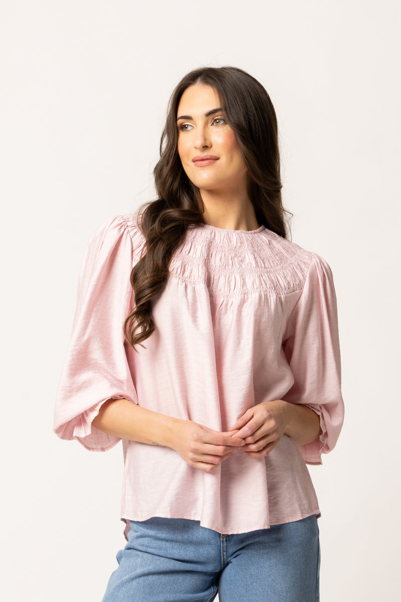 A woman with long dark wavy hair wearing a light pink smocked blouse with puff sleeves and blue jeans, standing against a plain white studio background.