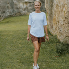 A smiling woman walking outdoors on a grassy area next to a large rock wall, wearing a light blue short-sleeve crewneck t-shirt, brown shorts, and white sneakers.