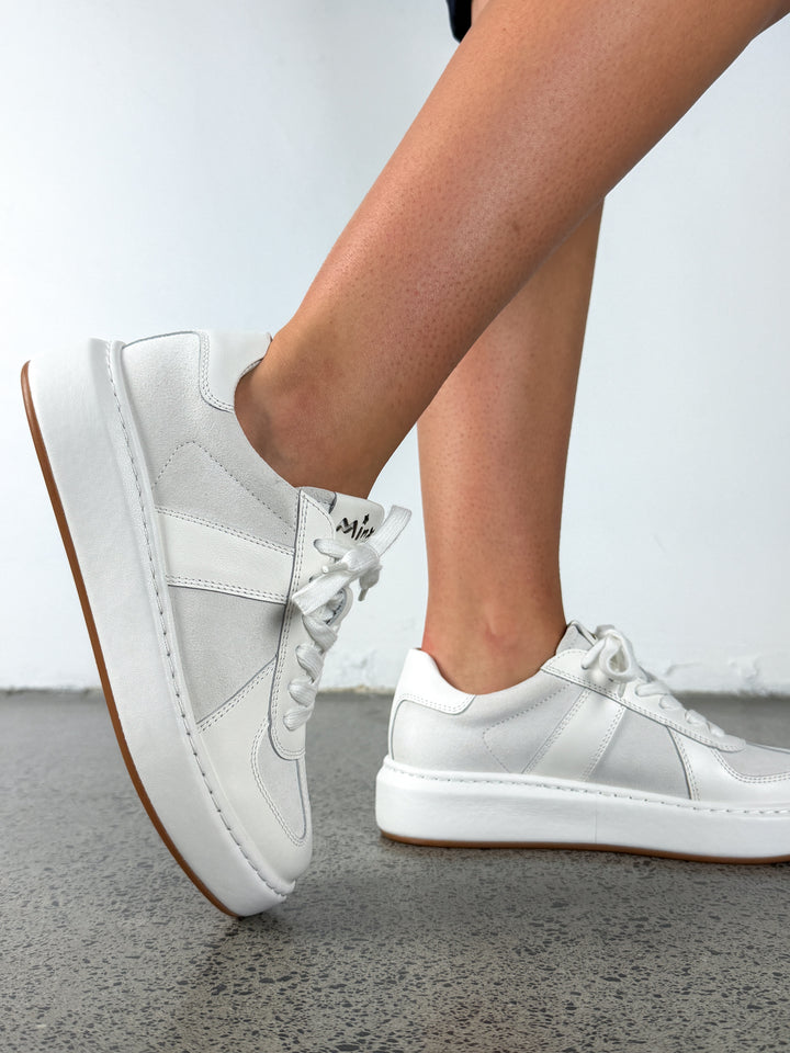 White platform sneakers with white laces and a gum sole, shown on a person's feet against a light gray wall and concrete floor.