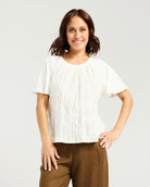 A woman modeling a white short-sleeve top with a textured crinkled fabric and a gathered neckline, paired with brown pants, against a light gray studio background.