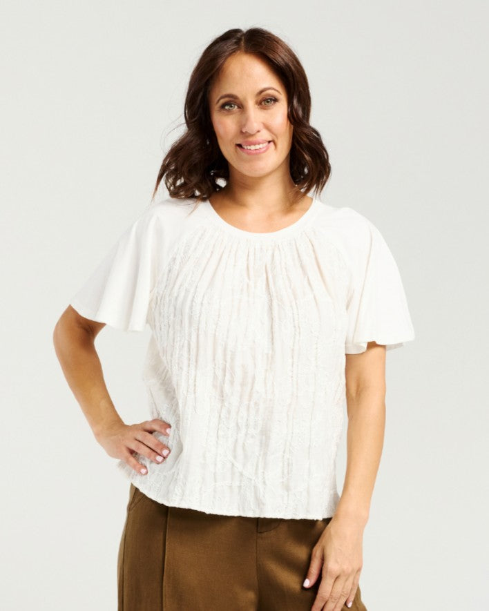 A woman modeling a white short-sleeve blouse with a textured crinkle fabric and a pleated round neckline, paired with brown pants, against a light gray studio background.