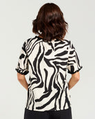 A woman viewed from the back wearing a short-sleeve blouse with a bold black and off-white zebra-like abstract pattern and ruffled cuffs, paired with dark bottoms, against a light gray studio background.