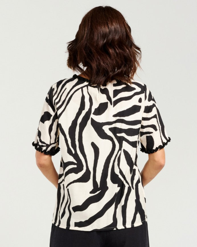 A woman viewed from the back wearing a short-sleeve blouse with a bold black and off-white zebra-like abstract pattern and ruffled cuffs, paired with dark bottoms, against a light gray studio background.