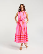 A woman modeling a sleeveless pink midi dress with vertical orange stripes, a matching tie belt at the waist, and horizontal orange stripes along the tiered hem, photographed against a light gray studio background.