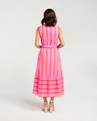 A woman modeling a sleeveless pink midi dress with vertical orange stripes, a matching tie belt at the waist, and horizontal orange stripes along the tiered hem, photographed against a light gray studio background.
