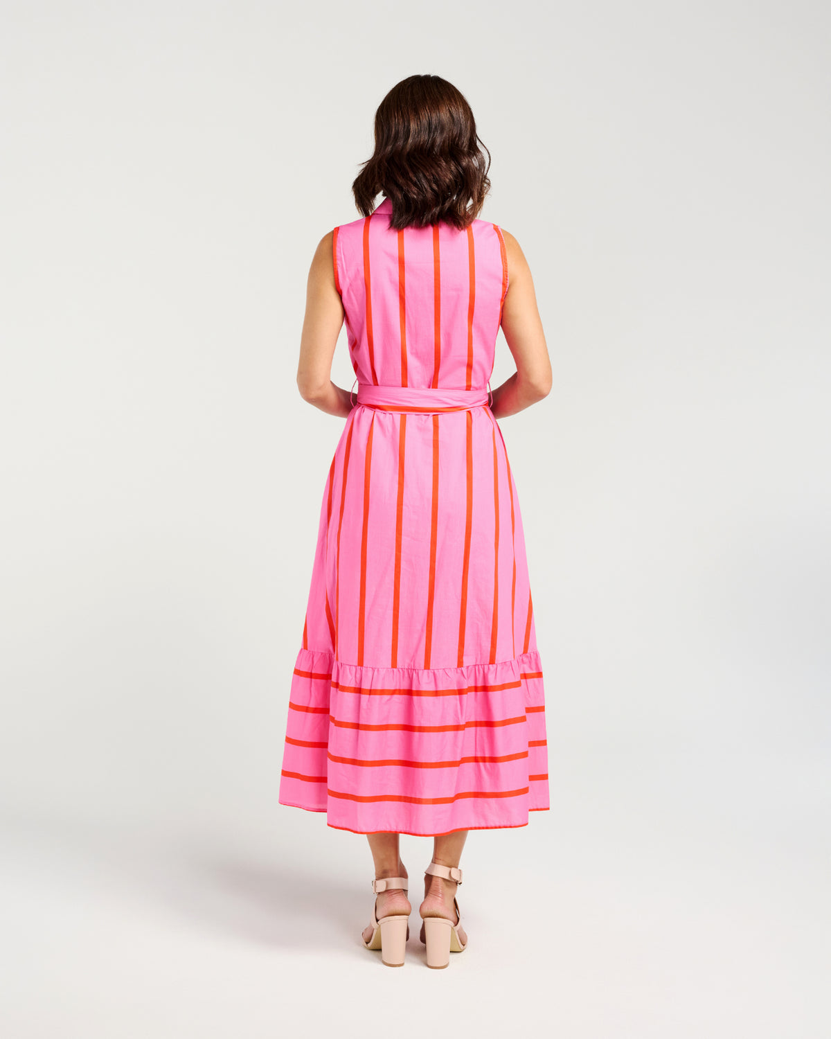 A woman modeling a sleeveless pink midi dress with vertical orange stripes, a matching tie belt at the waist, and horizontal orange stripes along the tiered hem, photographed against a light gray studio background.