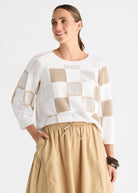 A woman wearing a white and beige checkered knit sweater with three-quarter sleeves and a beige skirt, photographed against a light gray studio background.