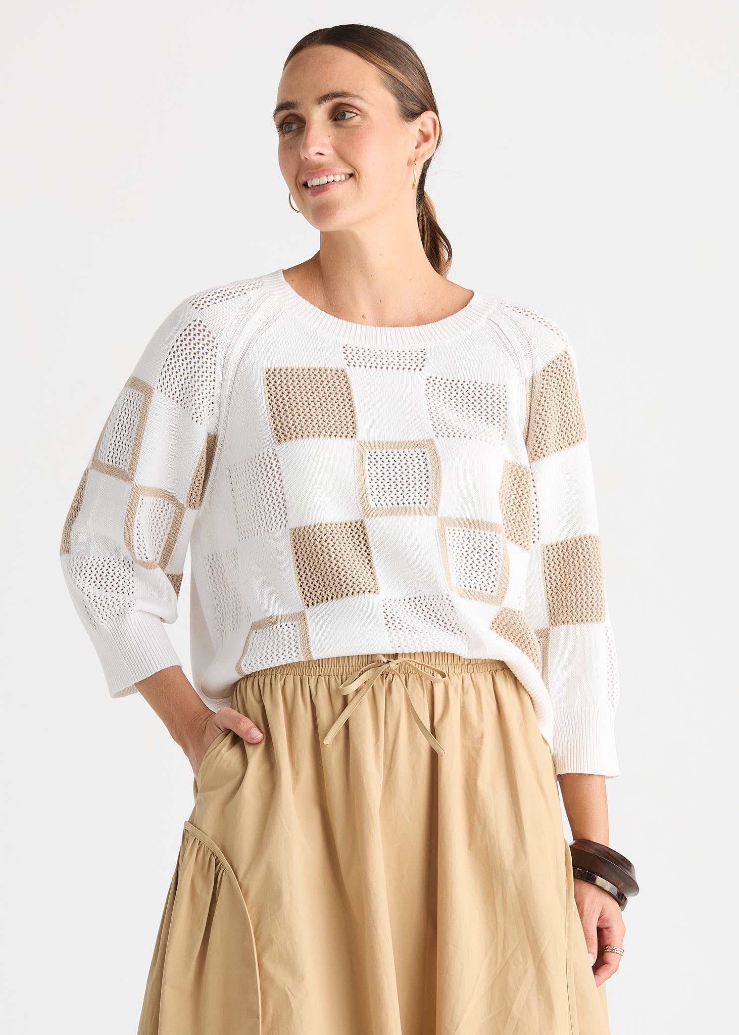 A woman wearing a white and beige checkered knit sweater with three-quarter sleeves and a beige skirt, photographed against a light gray studio background.
