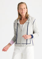 A woman wearing a light gray zip-up hoodie with navy blue contrast piping and cuffs, styled with white pants against a plain white studio background.