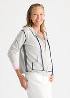 A smiling woman wearing a light gray zip-up hoodie with navy blue piping and stripes, paired with white pants, photographed against a clean white studio background.