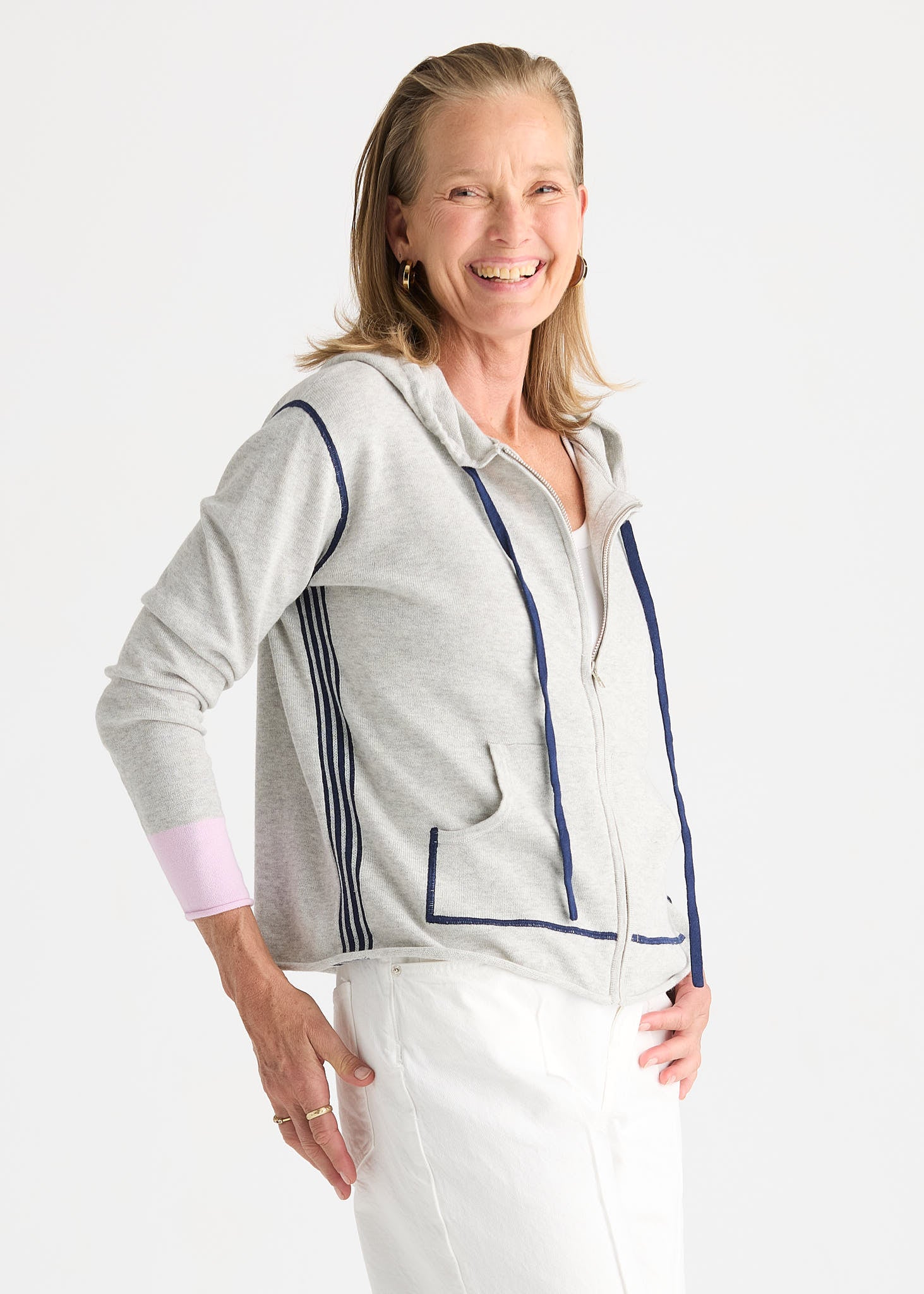 A smiling woman wearing a light gray zip-up hoodie with navy blue piping and stripes, paired with white pants, photographed against a clean white studio background.