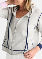 A woman wearing a light gray zip-up hoodie with navy blue contrast piping and drawstrings, layered over a white tank top.