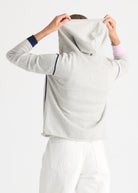 A person shown from the back wearing a light heather gray hooded sweatshirt with navy blue stripe accents on the sleeves and side, paired with white pants, against a plain white studio background.