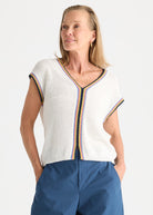 A woman wearing a white knit short-sleeve top with colorful striped trim and blue pants, photographed against a plain light background.